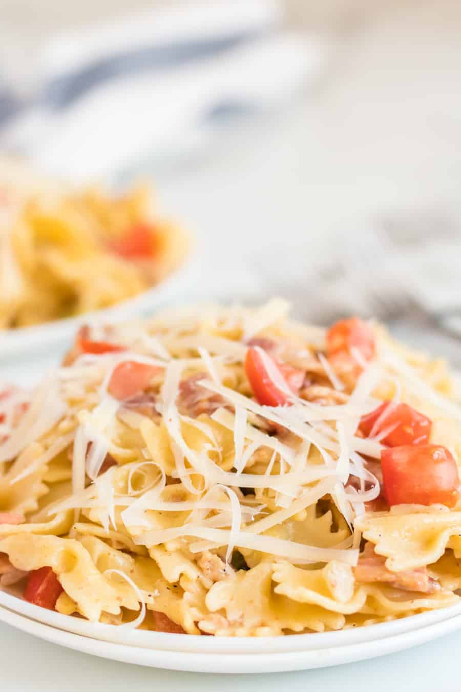 Pasta with bacon, tomatoes, green peppers, and shaved Parmesan cheese on a white plate with a fork.