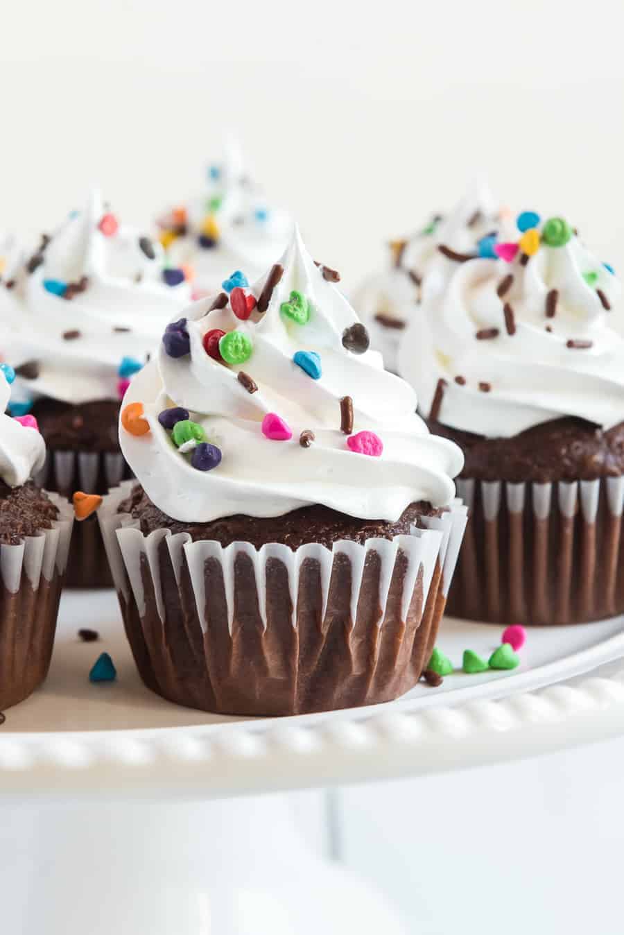 Chocolate cupcakes topped with swirls of white frosting without butter and colorful sprinkles are arranged on a white cake stand.