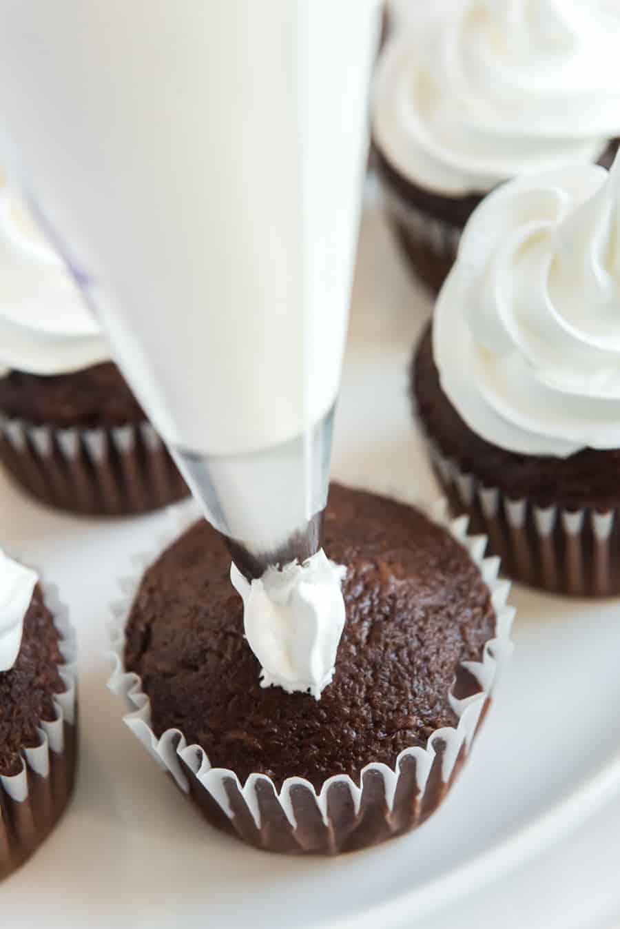 A close-up of a piping bag swirling white frosting without butter onto a chocolate cupcake, with other frosted cupcakes on a plate in the background.