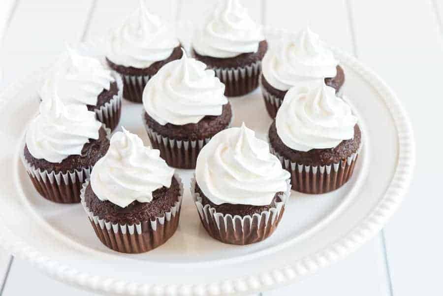 A white plate holds nine chocolate cupcakes, each topped with a swirl of white frosting without butter, set on a white surface.