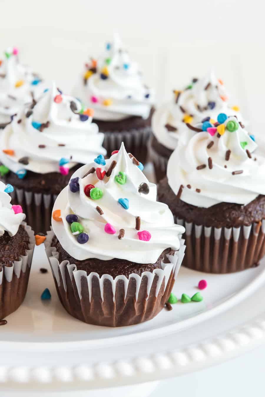 A plate of chocolate cupcakes with white swirled frosting without butter, topped with colorful sprinkles and chocolate shavings, displayed on a white surface.