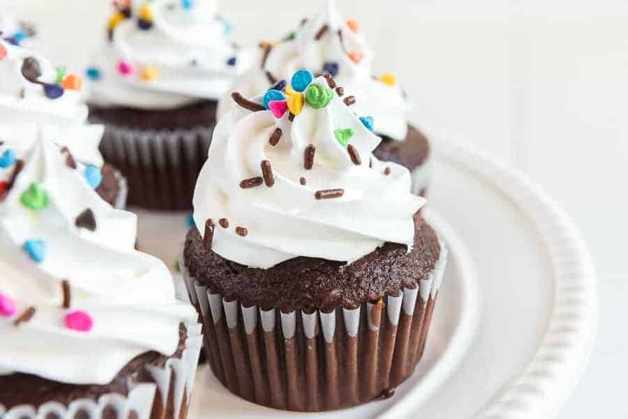 Chocolate cupcakes topped with swirls of frosting without butter, colorful round sprinkles, and chocolate sprinkles, arranged on a white plate.