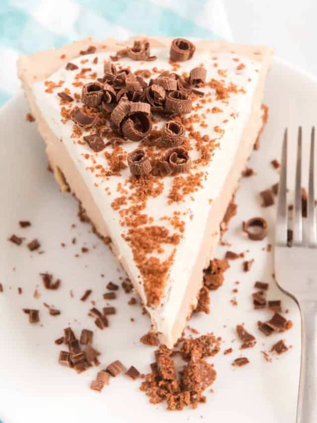A slice of creamy pie on a white plate, topped with chocolate shavings and crumbs. A fork is placed beside the slice, near the edge of the plate. The background features a blurred checkered pattern.