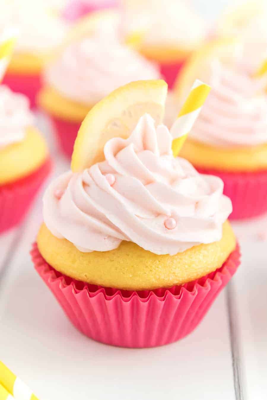 Pink Lemonade Cupcakes in bright pink wrappers with light pink frosting, pink sprinkles, a lemon slice, and a yellow and white straw.