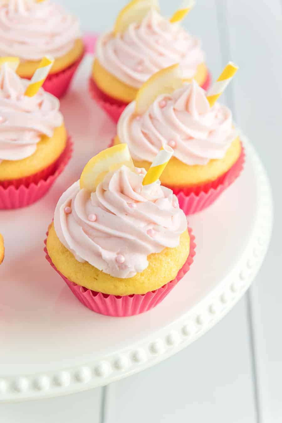 yellow cupcakes in bright pink wrappers with light pink frosting, pink sprinkles, a lemon slice, and a yellow and white straw on a white round serving platter.