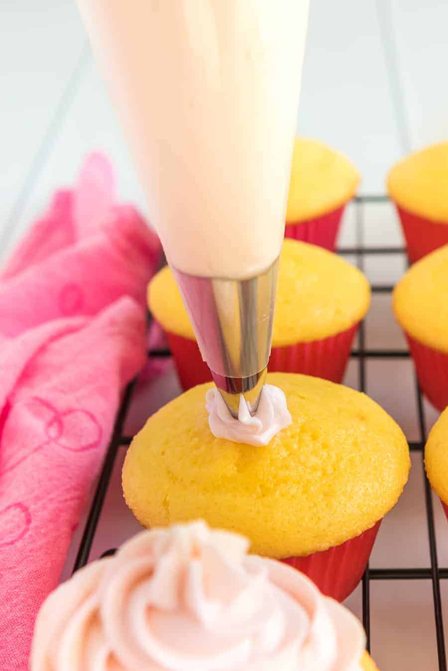 yellow cupcakes in bright pink wrappers with pale pink icing being piped on top.
