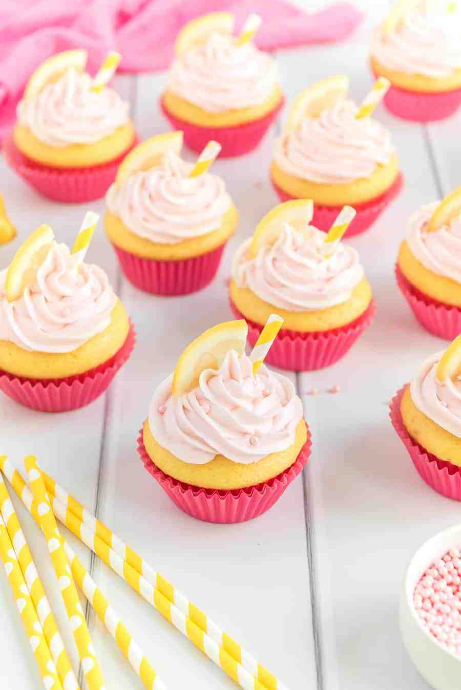 yellow cupcakes in bright pink wrappers with light pink frosting, pink sprinkles, a lemon slice, and a yellow and white straw on a white table.