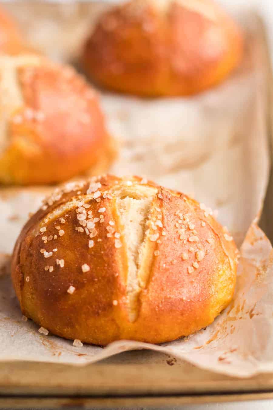 A closeup shot of a pretzel roll topped with coarse salt on a sheet pan lined with parchment paper.