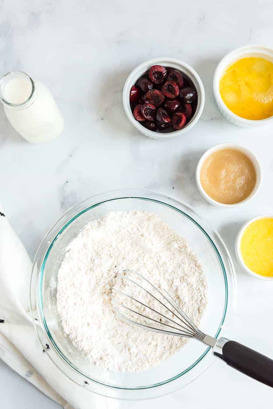A bowl of flour with a whisk sits on a marble countertop, ready to craft cherry waffles. Nearby are small bowls containing cherries, mashed bananas, a golden liquid, and a glass milk bottle. A white cloth is partially visible on the left.