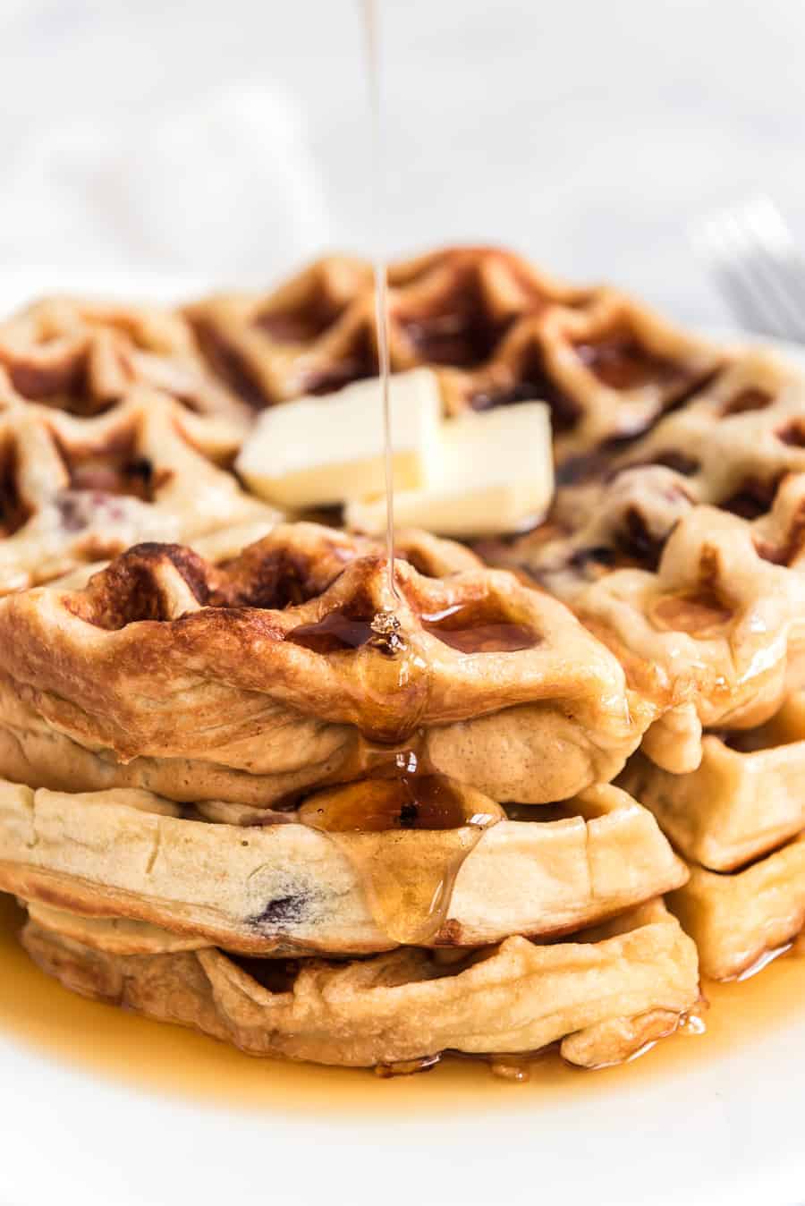 A stack of cherry waffles topped with a pat of butter is being drizzled with syrup. The waffles are golden brown and fluffy, sitting on a white plate. The syrup glistens as it flows down the sides.