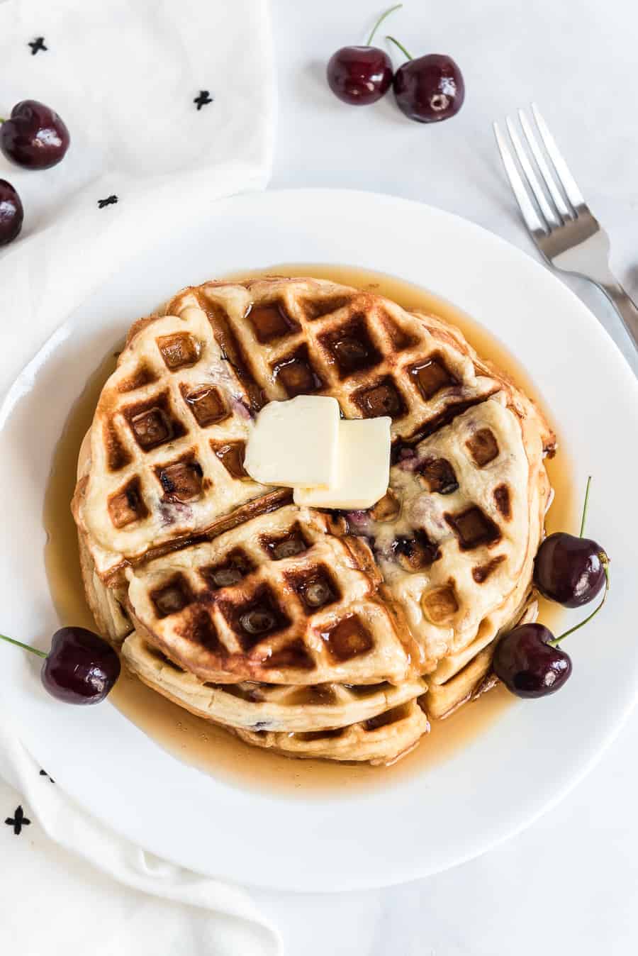 A plate of sweet cherry waffles topped with two pats of butter and drizzled with syrup. Fresh cherries are artfully scattered around the plate, adding a pop of color. A fork rests beside it on a crisp white cloth.