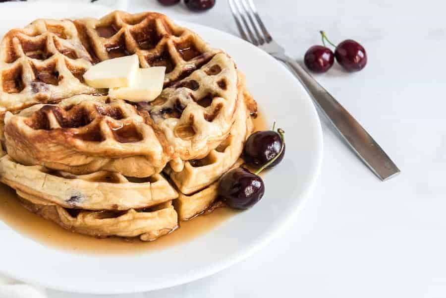 A stack of cherry waffles topped with melting butter and drizzled with syrup sits on a white plate. Fresh cherries are scattered around, enhancing the delightful presentation. A fork is placed to the side, ready for a delicious dive.