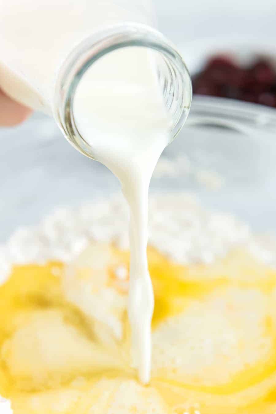 Milk cascades from a glass bottle into a bowl, mingling with flour and a cracked egg. In the softly blurred background, the faint outlines of dark-colored fruit hint at delicious cherry waffles ready to be created.