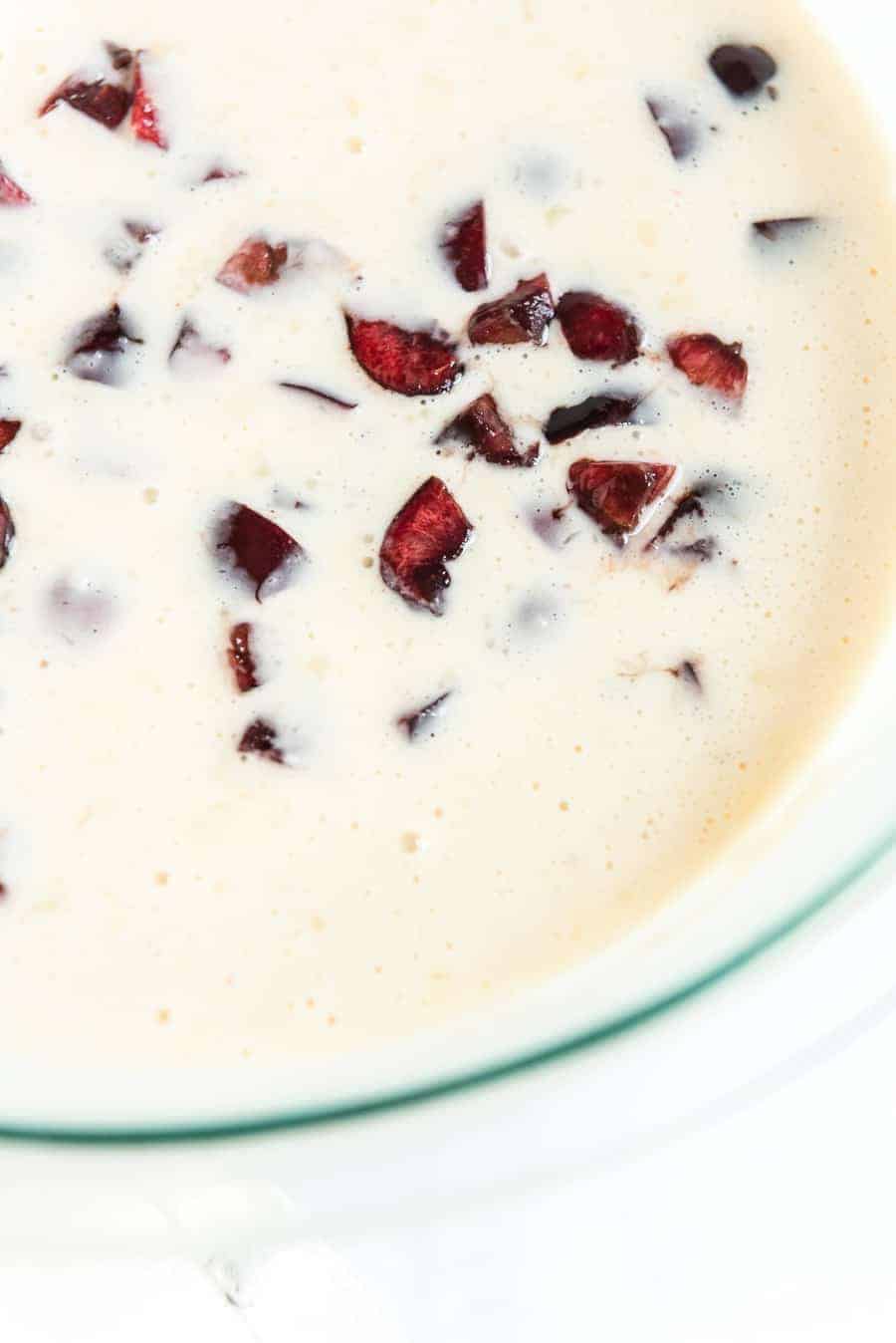 Close-up of a creamy mixture with small chunks of dark red fruit, likely cherries, scattered throughout. The cream appears smooth and slightly frothy, reminiscent of the toppings on cherry waffles, creating a contrast with the shiny fruit pieces.