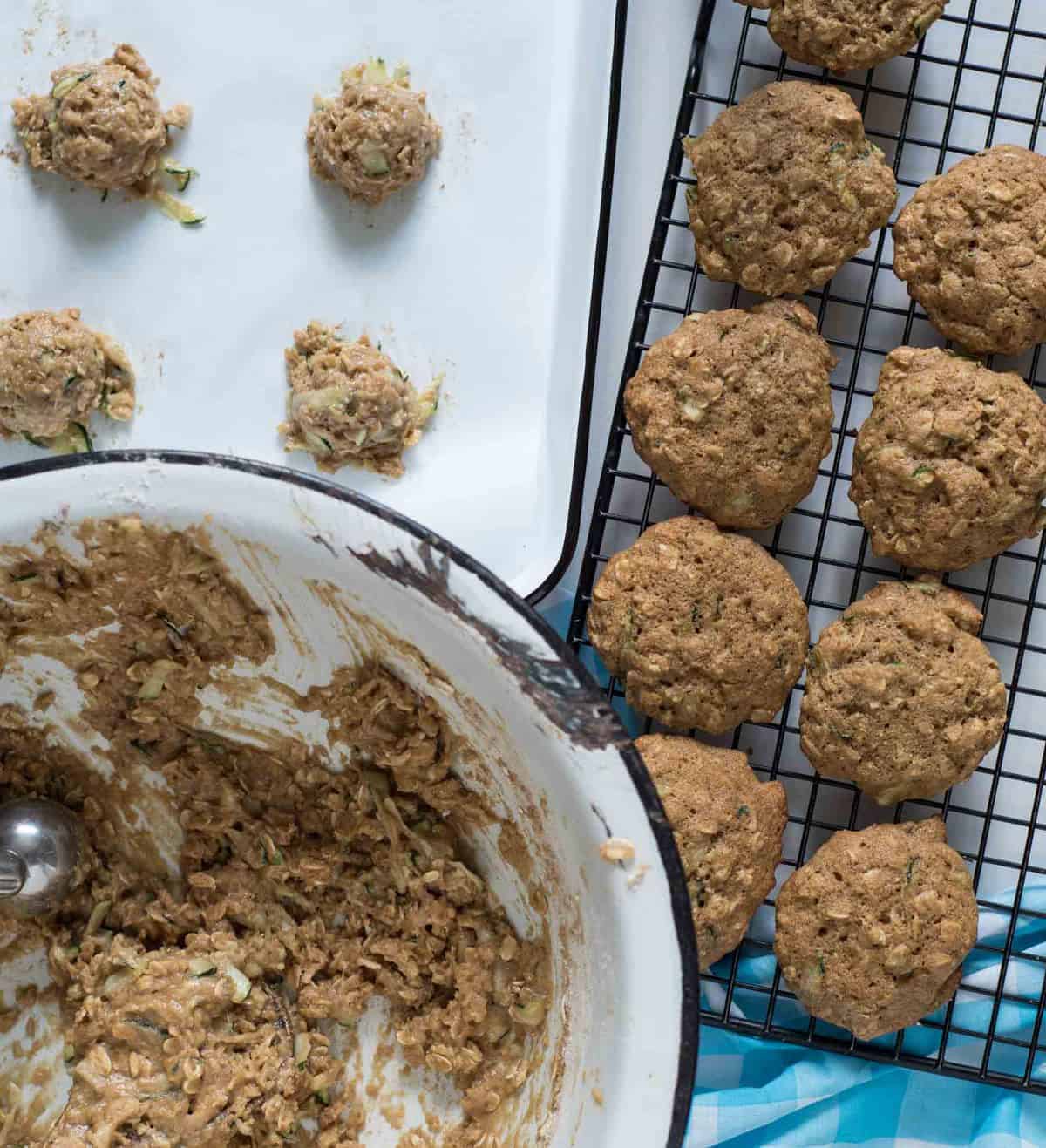 A dozen baked zucchini oatmeal cookies on a black wire cooling rack next to the bowl of batter and a baking sheet with four scoops of batter about to be baked.