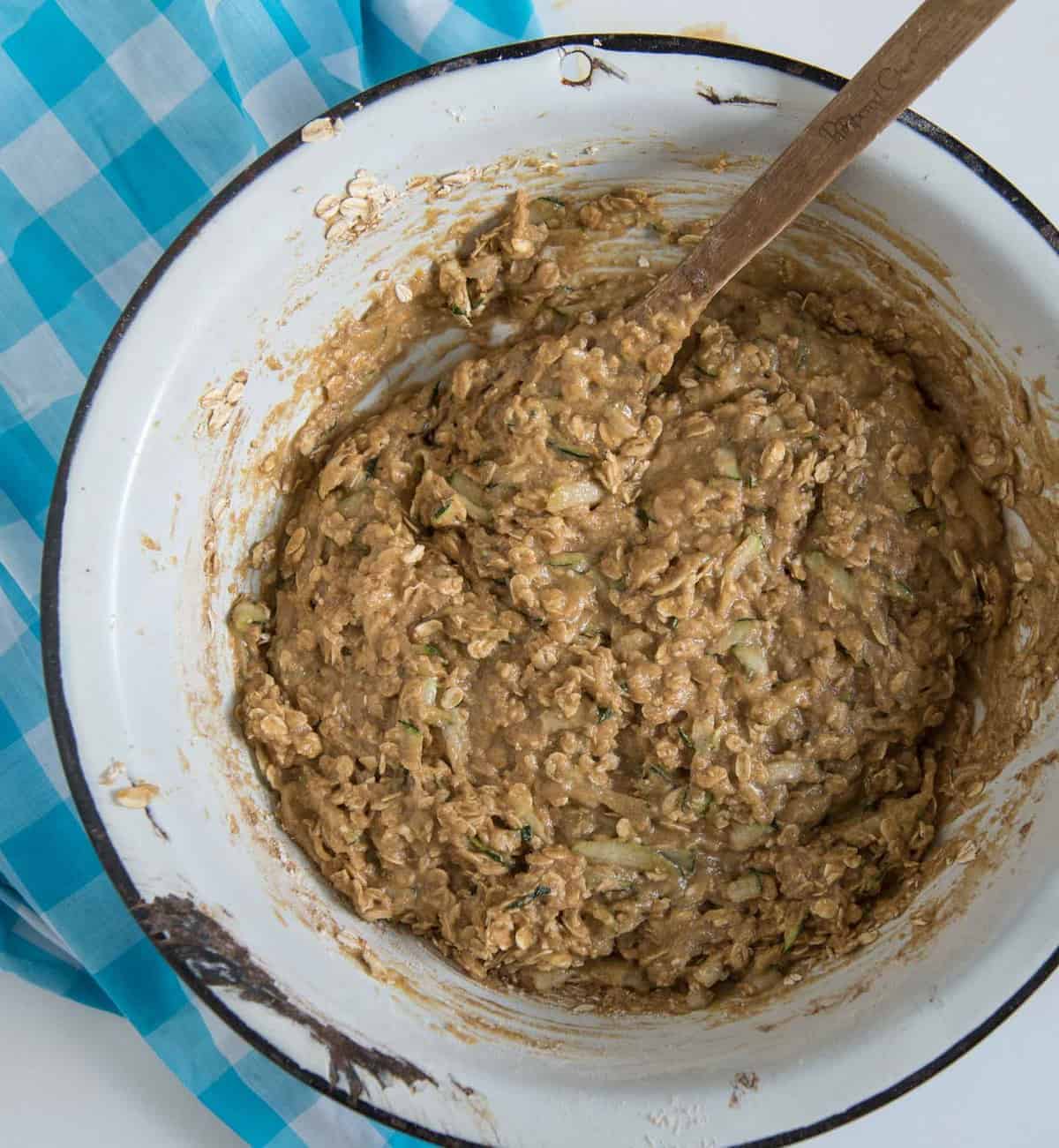 The batter for zucchini oatmeal cookies being mixed with a wooden spoon in a white enamelware bowl with a black rim.