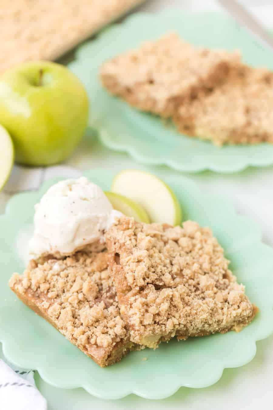 Two crumbly apple dessert bars, reminiscent of apple crumb cake, sit on a green plate with vanilla ice cream and apple slices. Another plate with more bars is in the background, alongside a whole green apple.