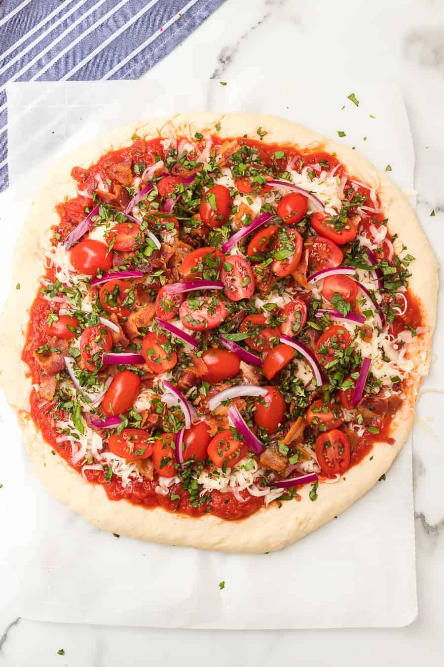 unbaked pizza dough with sauce, mozzarella, red tomatoes, green herbs, and red onion toppings on a white parchment.