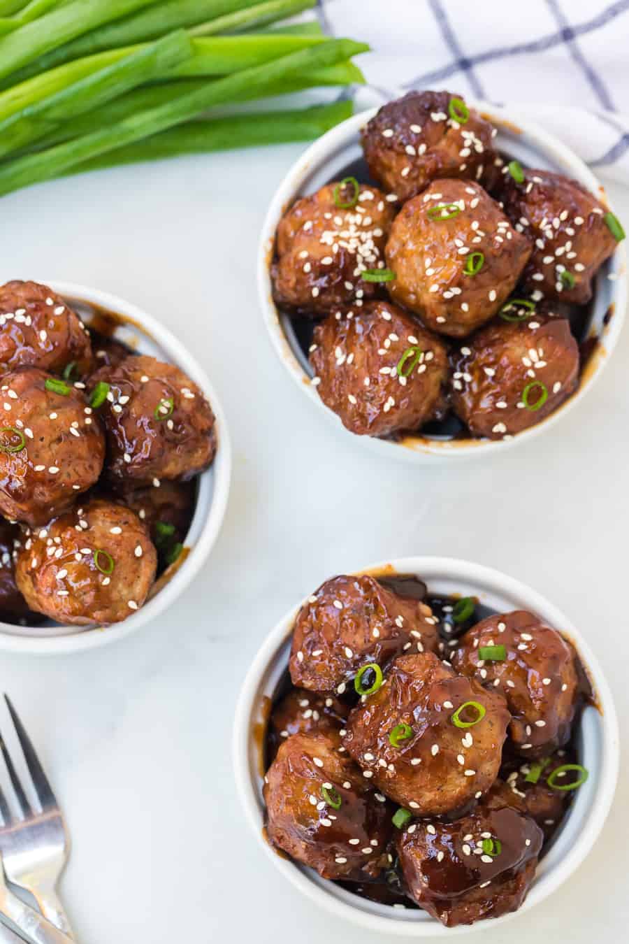three white ramekins full of sweet savory meatballs topped with sesame seeds and green onions.