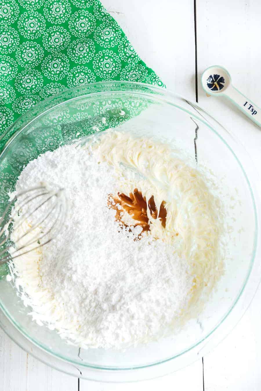 A glass bowl with flour in it as well as a splash of vanilla extract. A beater is on the edge of the bowl and a measuring spoon is lying near by.