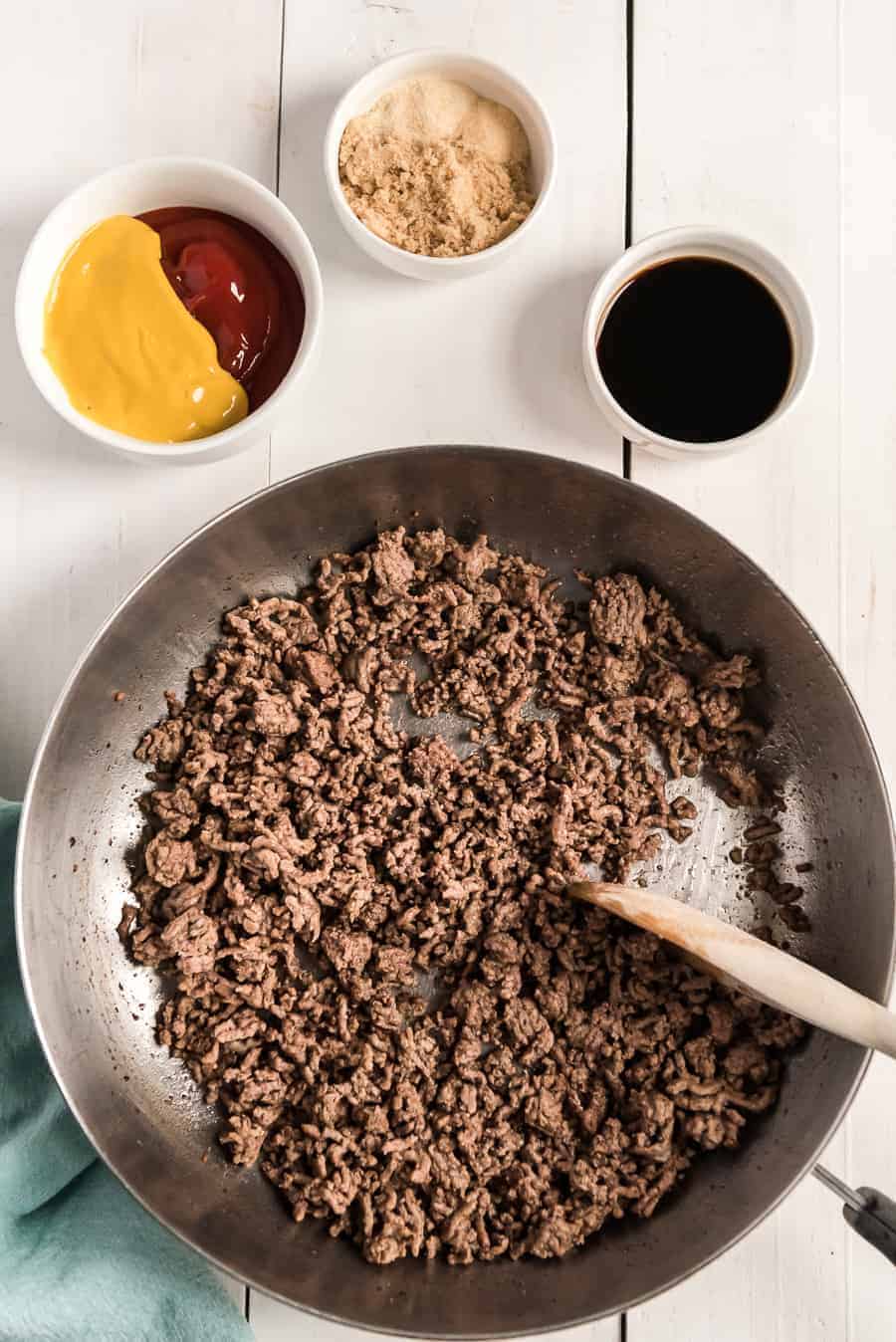 a metal pan with ground beef a white bowl with mustard and ketchup white bowl with dark liquid smaller white bowl with brown sugar.