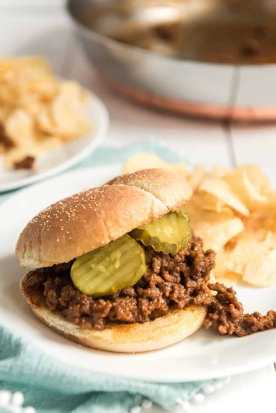 white plate containing sloppy joe on a bun with two pickles and potato chips.