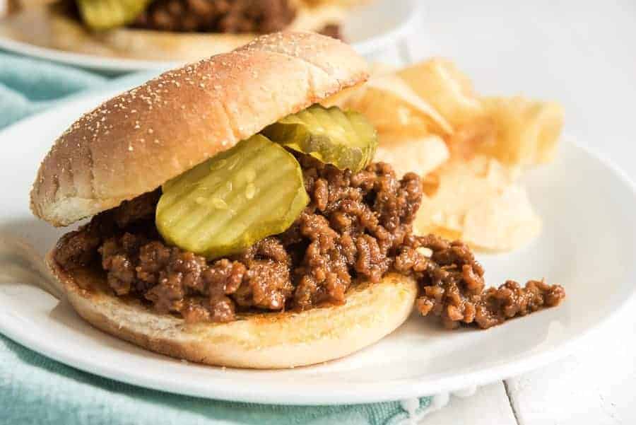 white plate containing sloppy joe on a bun with two pickles and potato chips.