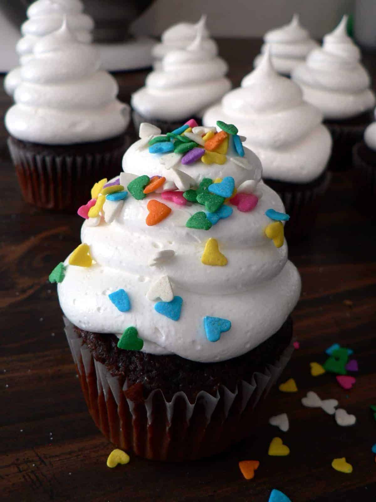 Chocolate cupcake with swirls of white frosting without butter, topped with colorful heart-shaped sprinkles, sits on a dark wooden surface. Other similarly decorated cupcakes are in the background.