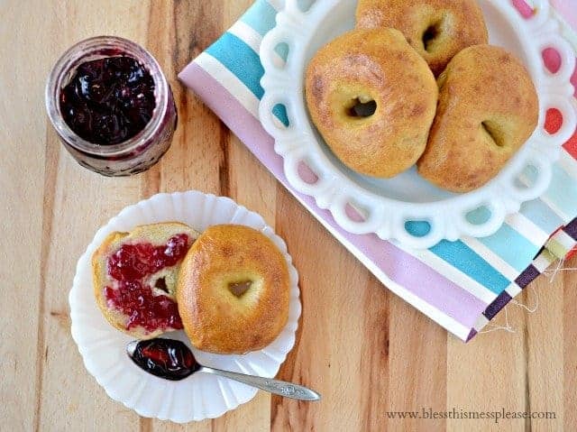 3 bagels on a plate in the top right corner. a jar of jam in the top left corner. a small plate with a bagel sliced in half, spread with jam and a spoon in the lower left.