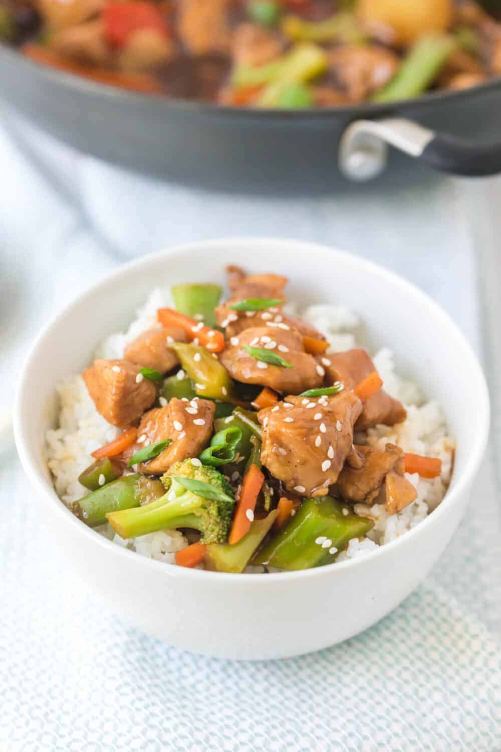 A white bowl filled with fluffy white rice, topped with chicken stir fry with vegetables—broccoli, carrots, and greens—garnished with sesame seeds. A blurred pan of similar food sits in the background.
