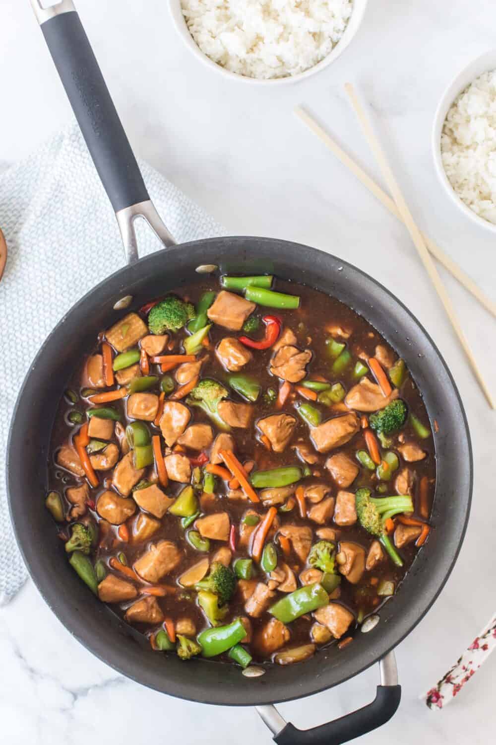 stir fry chicken and vegetables in a skillet next to chopsticks and bowls of white rice.