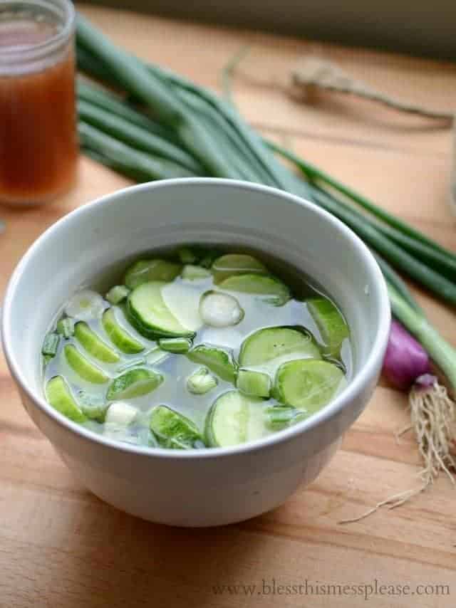 A white bowl filled with quick pickled cucumbers and green onion slices in clear liquid rests on a wooden surface. Fresh green onions and a glass jar are in the background.