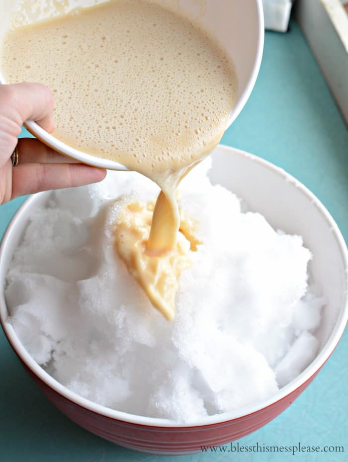 A hand pours creamy yellow liquid from a bowl over a large bowl of freshly gathered snow, starting a delicious Snow Ice Cream Recipe on a teal countertop.