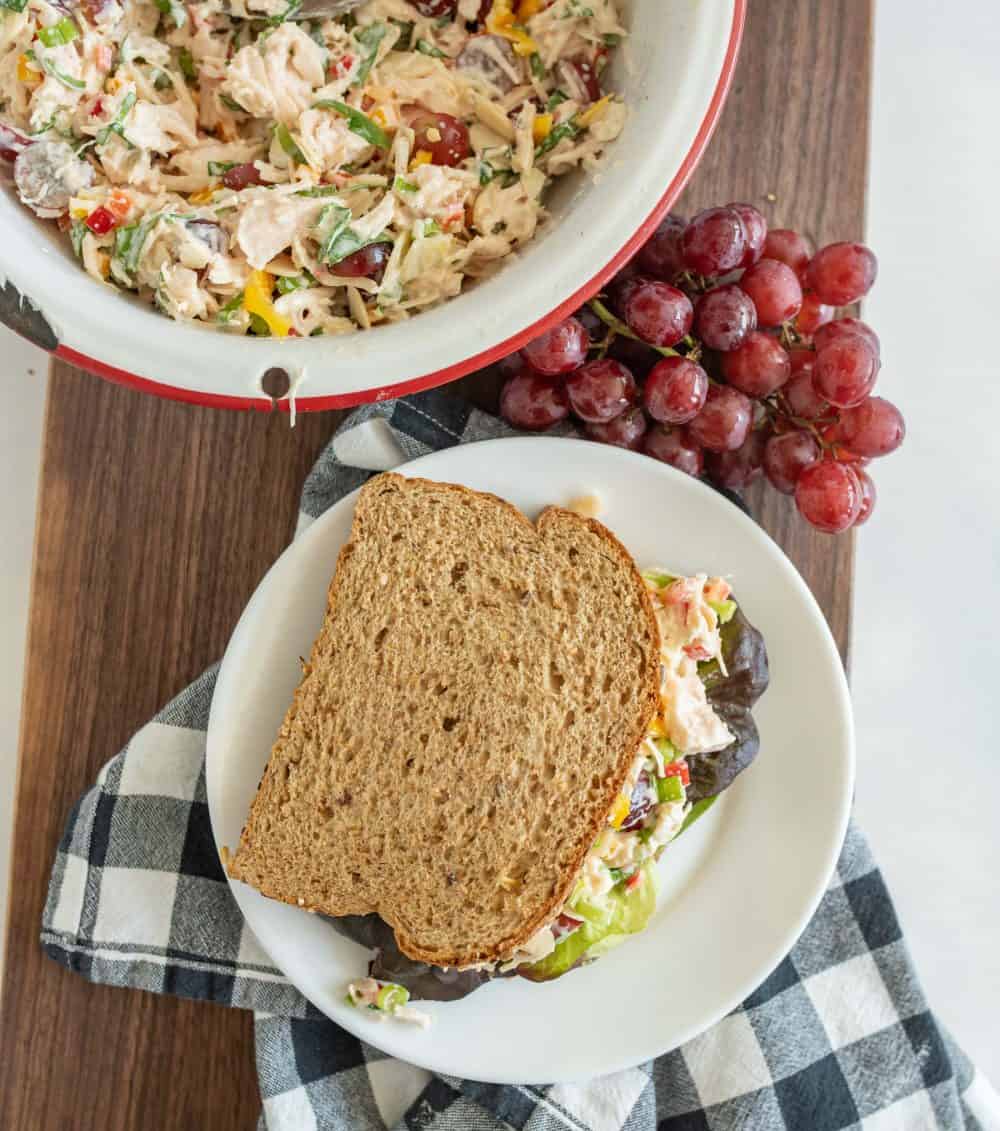 top view of a bowl of chicken salad, a bunch of grapes, and a chicken salad sandwich resting on a cutting board