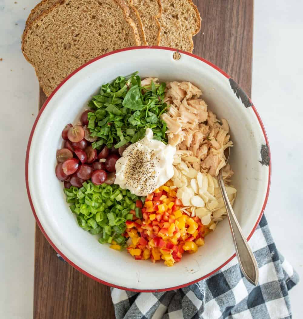 top view of a bowl with all the ingredients for chicken salad sitting on a cutting board with whole grain bread above it and a plaid dish towel below it