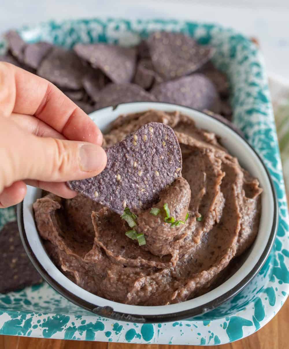 a hand dipping a blue corn tortilla chip in a bowl of black bean dip