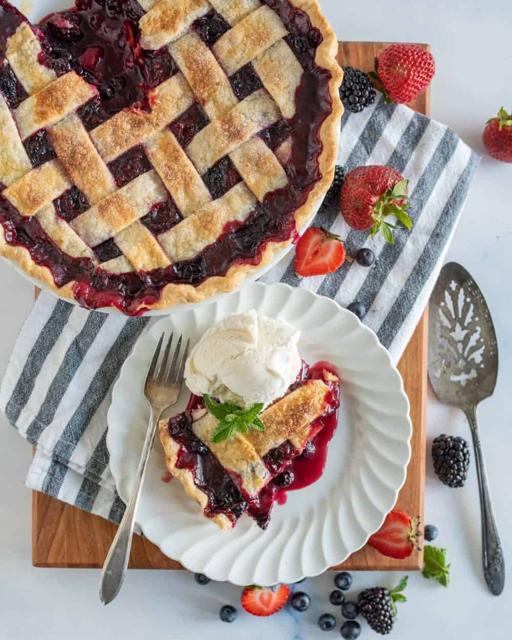 top view of a slice of triple berry pie on a plate next to the pie, serving utensils, and berries.