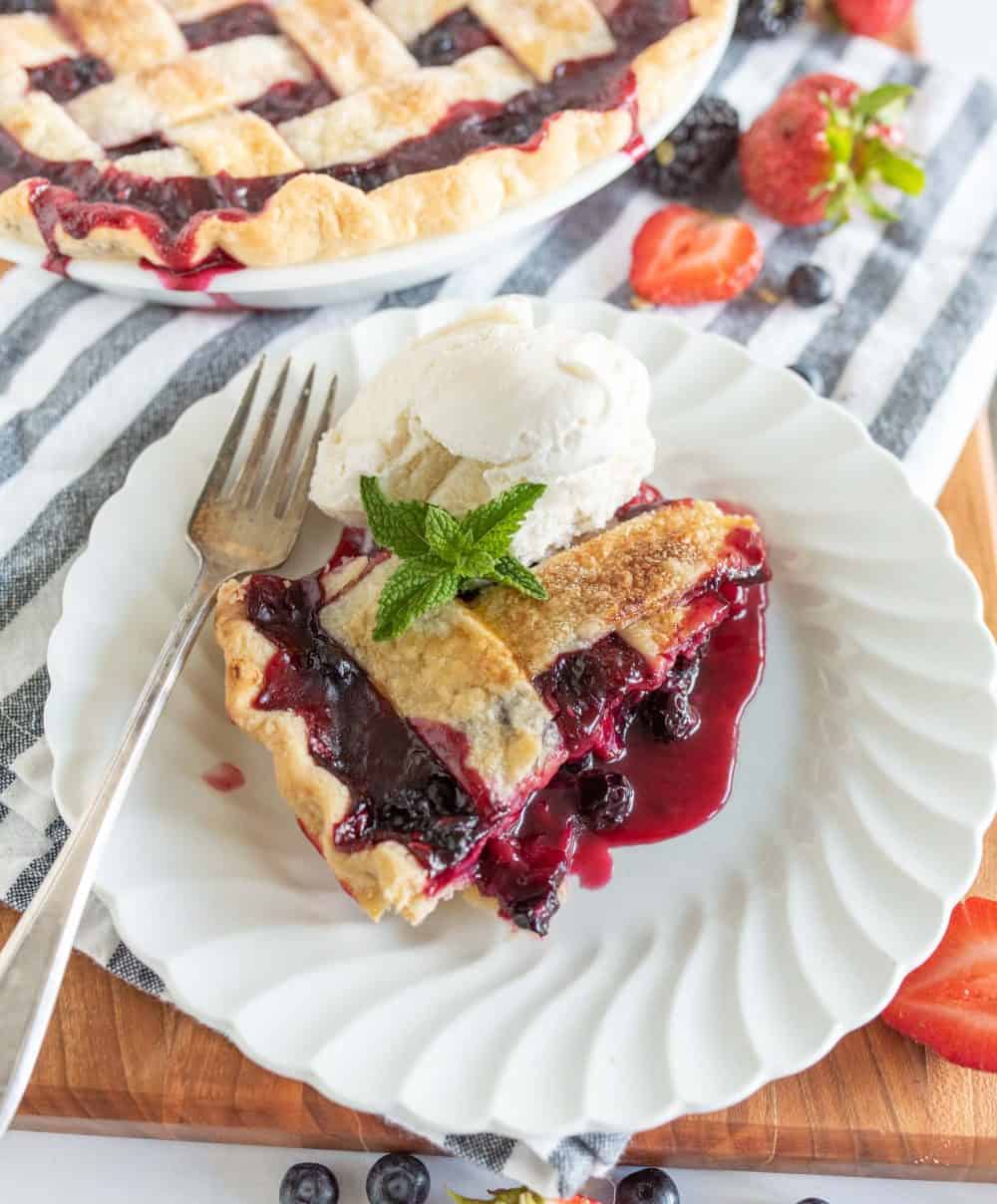 close up picture of a slice of triple berry pie on a plate with ice cream and a mint leaf.