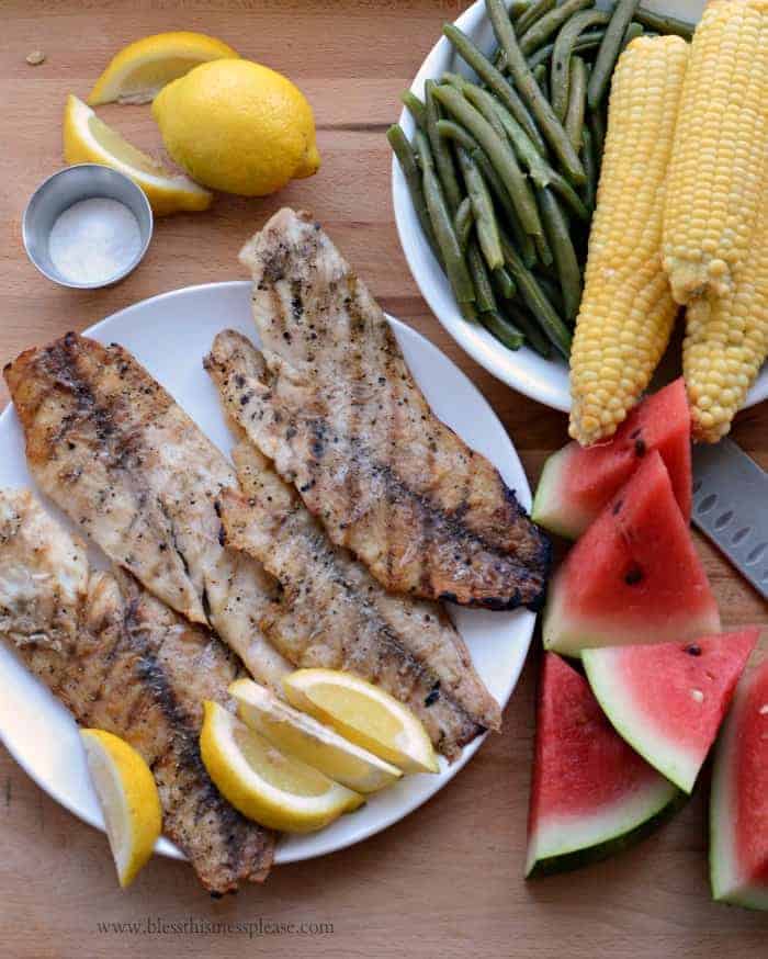 A plate with three grilling fish fillets and lemon wedges is surrounded by fresh green beans, corn on the cob, watermelon slices, a whole lemon, and a small dish of salt on a wooden surface.