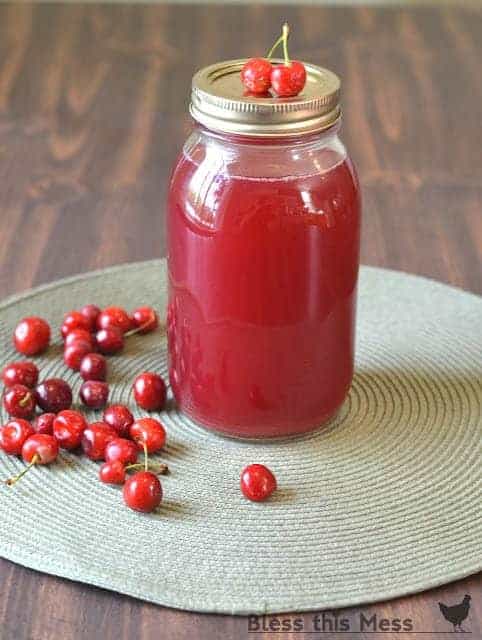 A jar of homemade juice with cherries on top sits on a round gray mat, showcasing a delightful cherry juice recipe. A small cluster of fresh cherries is scattered around the jar against a dark wooden background.