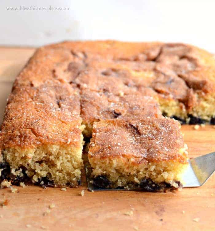 Close-up of a freshly baked cinnamon cake on a wooden surface. A spatula lifts one square piece from the cake, revealing a moist interior with a layer of berries at the bottom. The top is sprinkled with sugar, giving it a golden-brown crust.