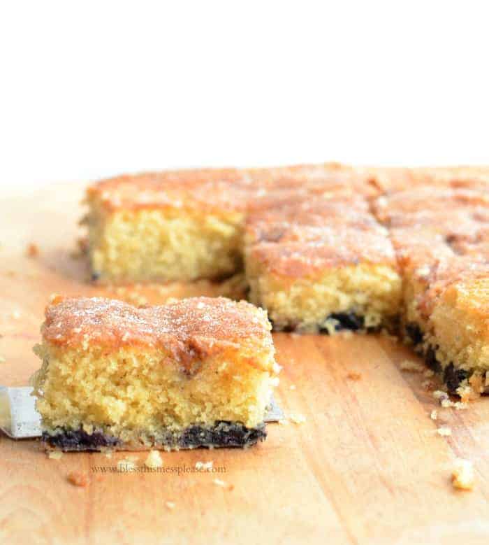 A close-up of square slices of coffee cake with a golden, crumbly top, set on a wooden board. One piece is being lifted with a spatula, revealing a dark, layered bottom crust.