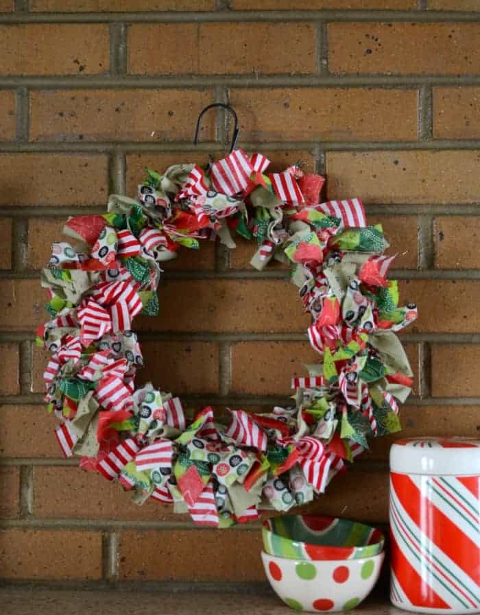 DIY Christmas Rag Wreath made from red, green, and white fabrics hanging on a brick wall with a couple Christmas bowls and a candy tin next to it.