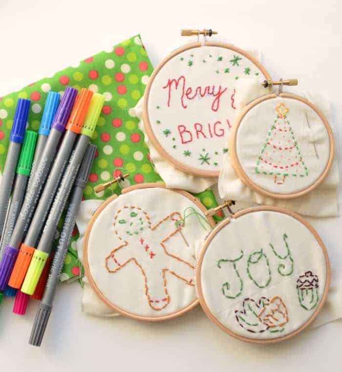 Four simple embroidered Christmas ornaments in hoops before finishing next to some green and red fabric and some washable fabric markers.
