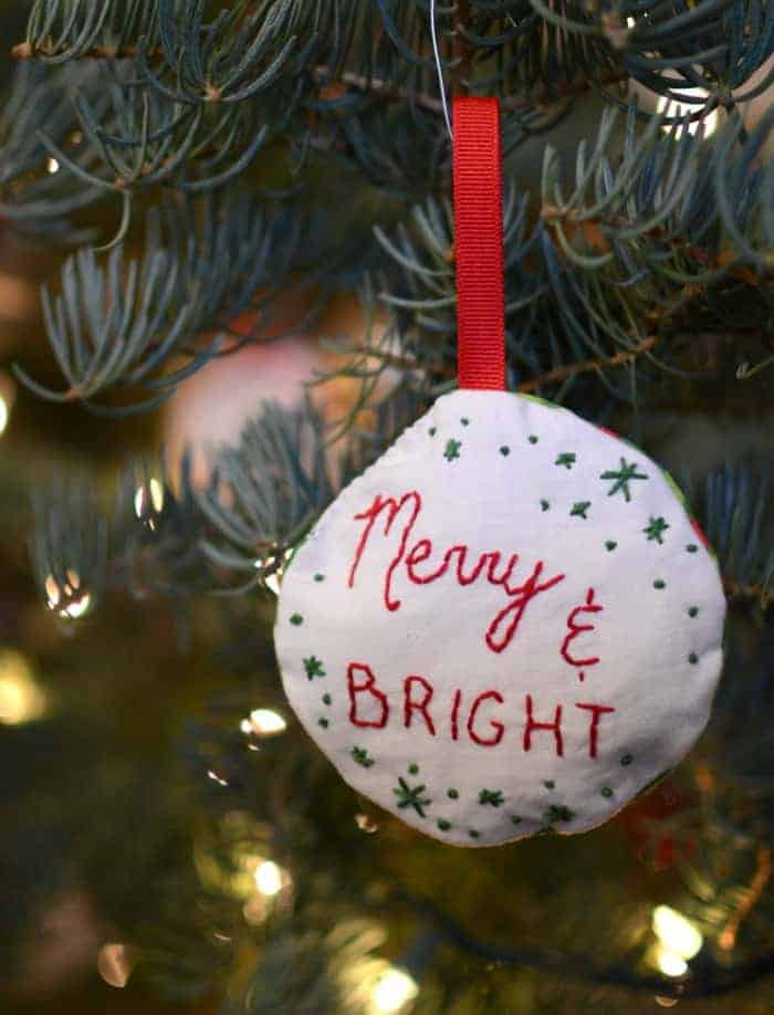Stuff embroidered ornament that says merry and bright hanging on a Christmas tree.