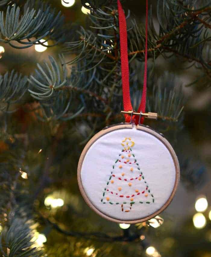 A simple embroidered ornament with a Christmas tree on it in a hoop hanging on a Christmas tree.