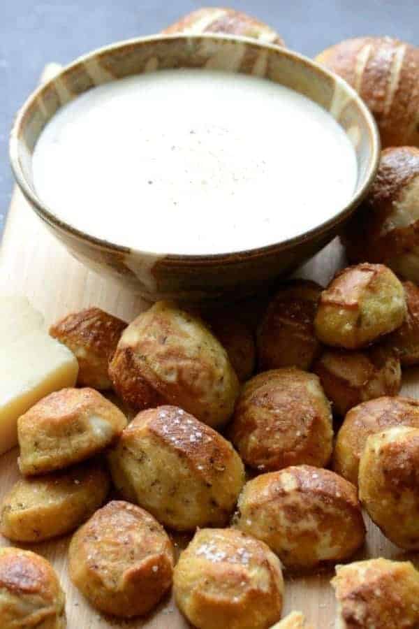 A close-up of the baked parmesan pretzel bites on a bamboo cutting board with a bowl of the parmesan dipping sauce.