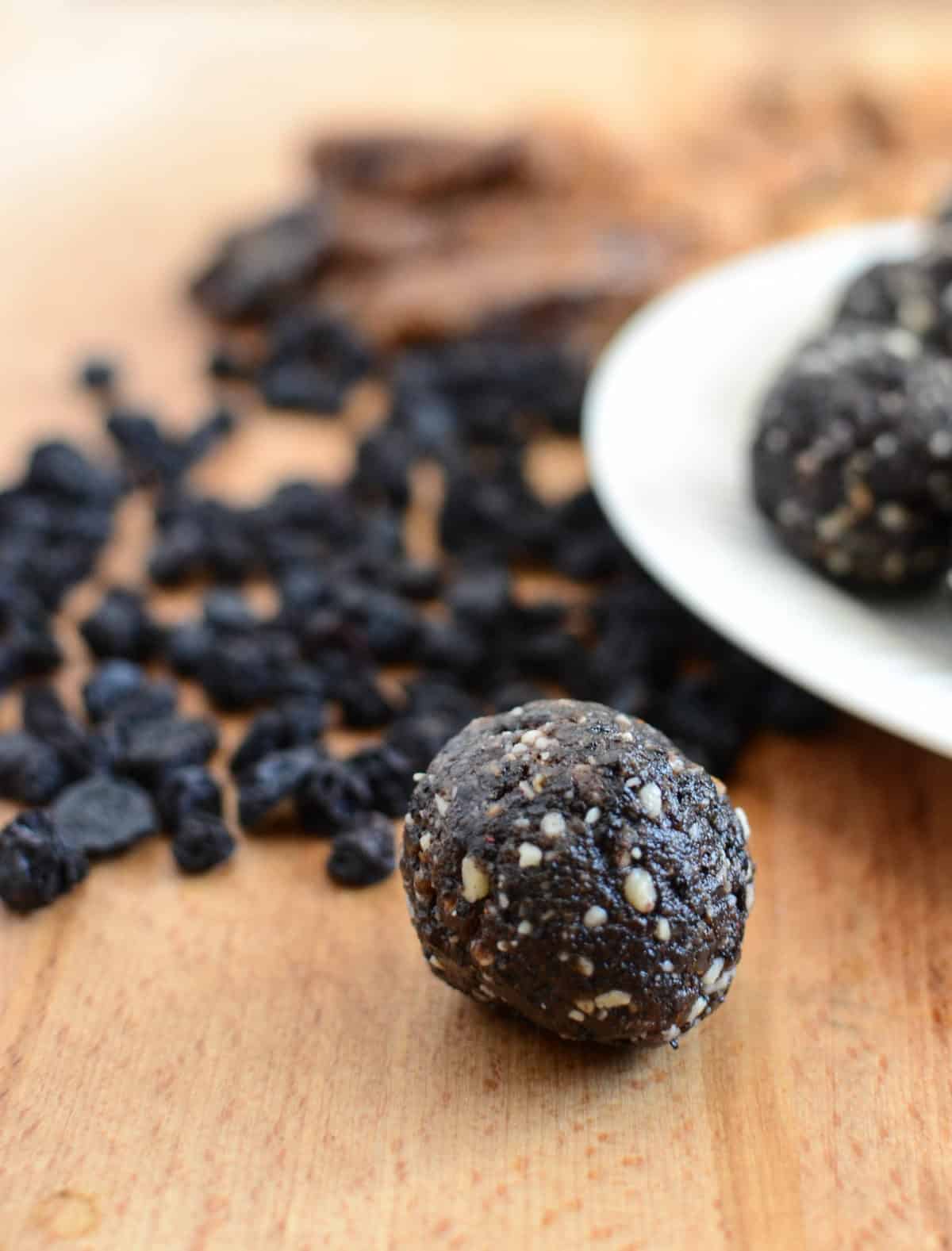 A close-up of a homemade energy ball on a wooden surface, showcasing visible nuts and seeds. In the background, a plate with more blueberry muffin date balls and scattered dried blueberries are partially visible.