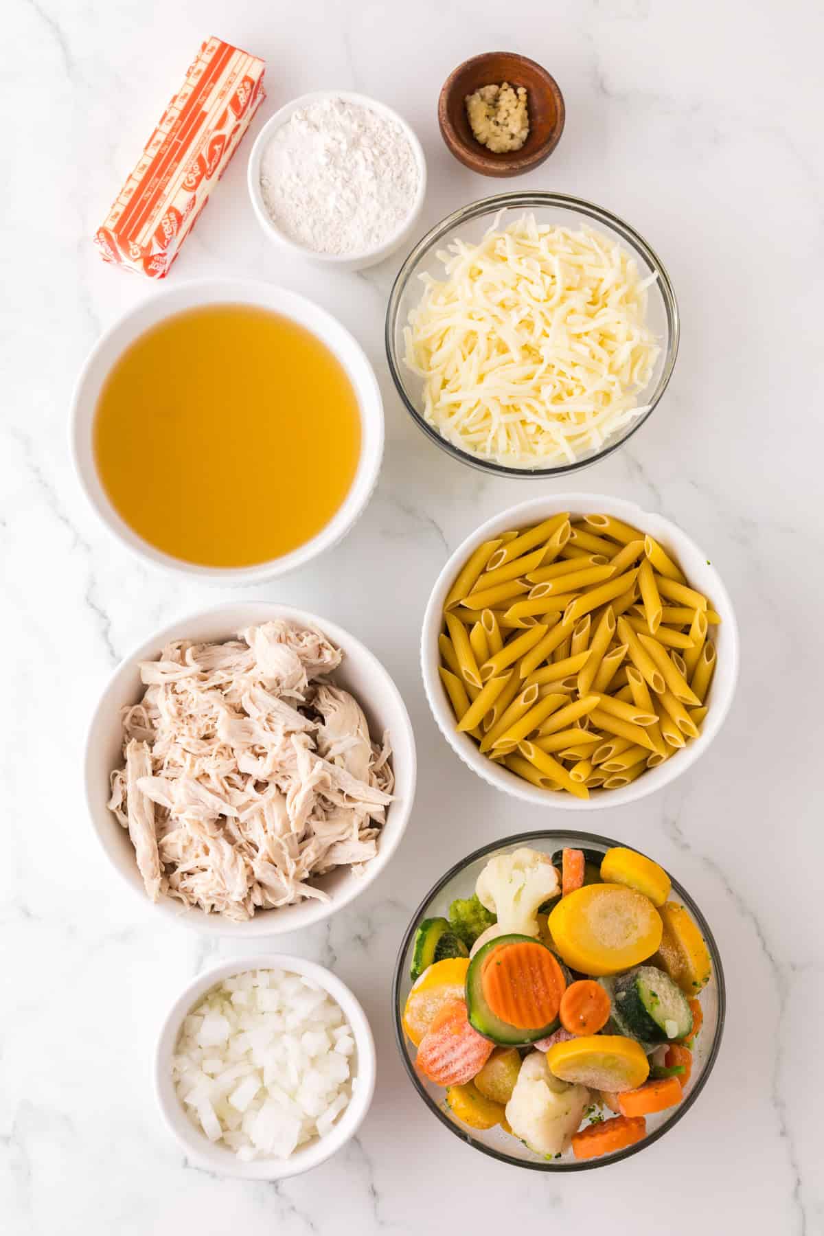 Overhead view of chicken vegetable pasta ingredients in bowls on a white surface: butter, flour, minced garlic, shredded cheese, broth, uncooked penne pasta, shredded cooked chicken, mixed vegetables, and chopped onions.