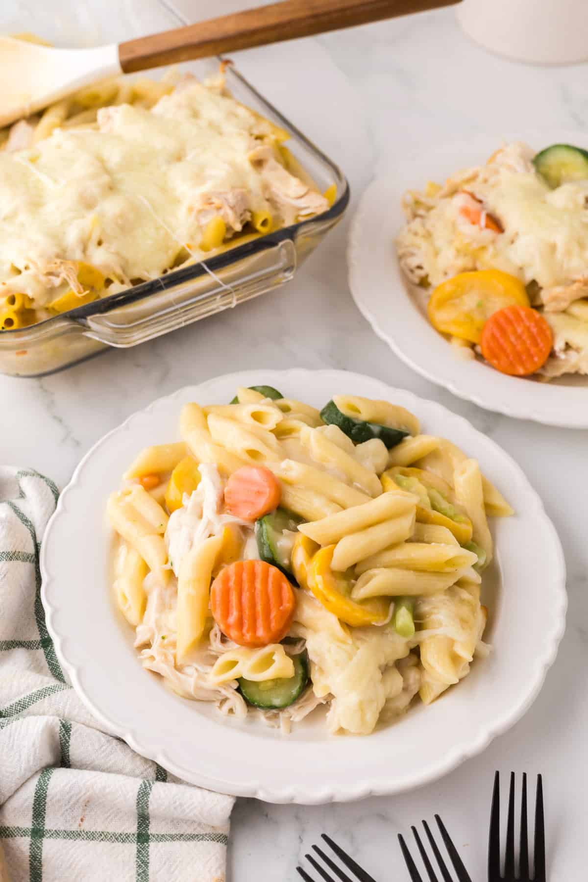 A plate of baked chicken vegetable pasta with sliced carrots, zucchini, and melted cheese, with a casserole dish of the same pasta in the background. A checkered towel and black forks are nearby.